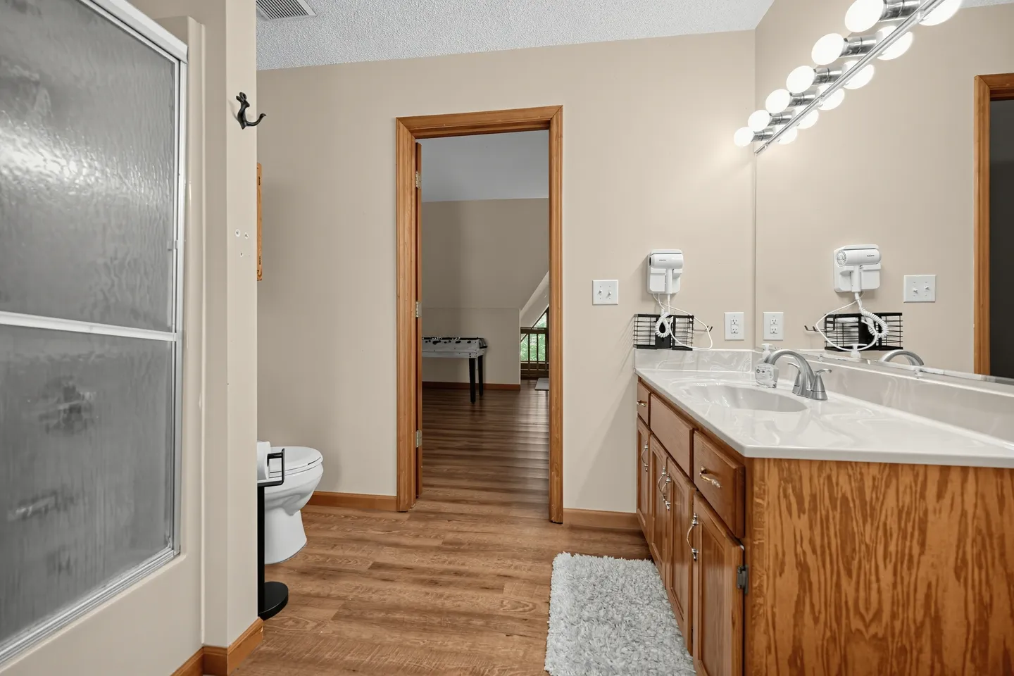 Bathroom featuring a wood vanity with a white countertop, vanity mirror lighting, a toilet, a textured glass shower door, and wood-look flooring looking into an adjacent room.
