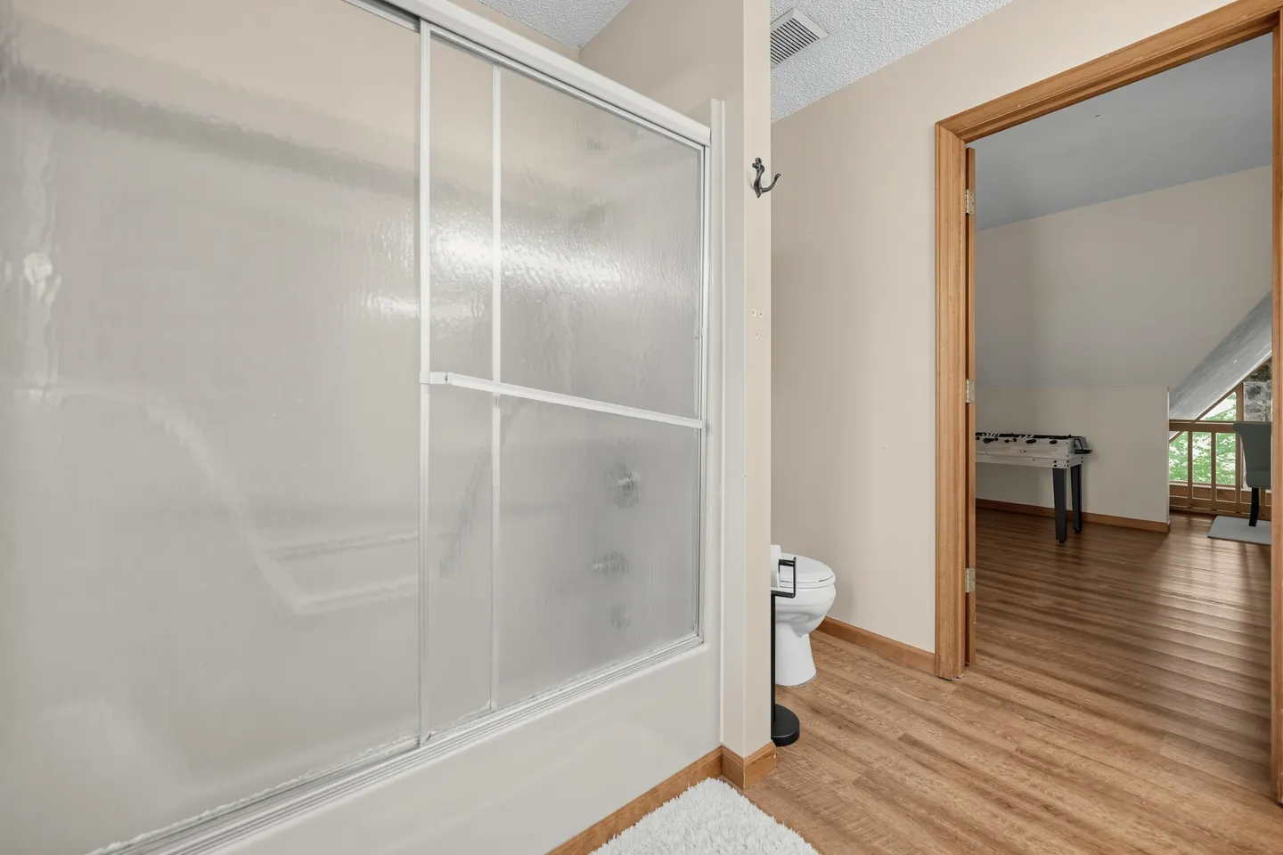Bathroom with a bathtub and sliding textured glass doors, next to a toilet and an open doorway leading to another room.