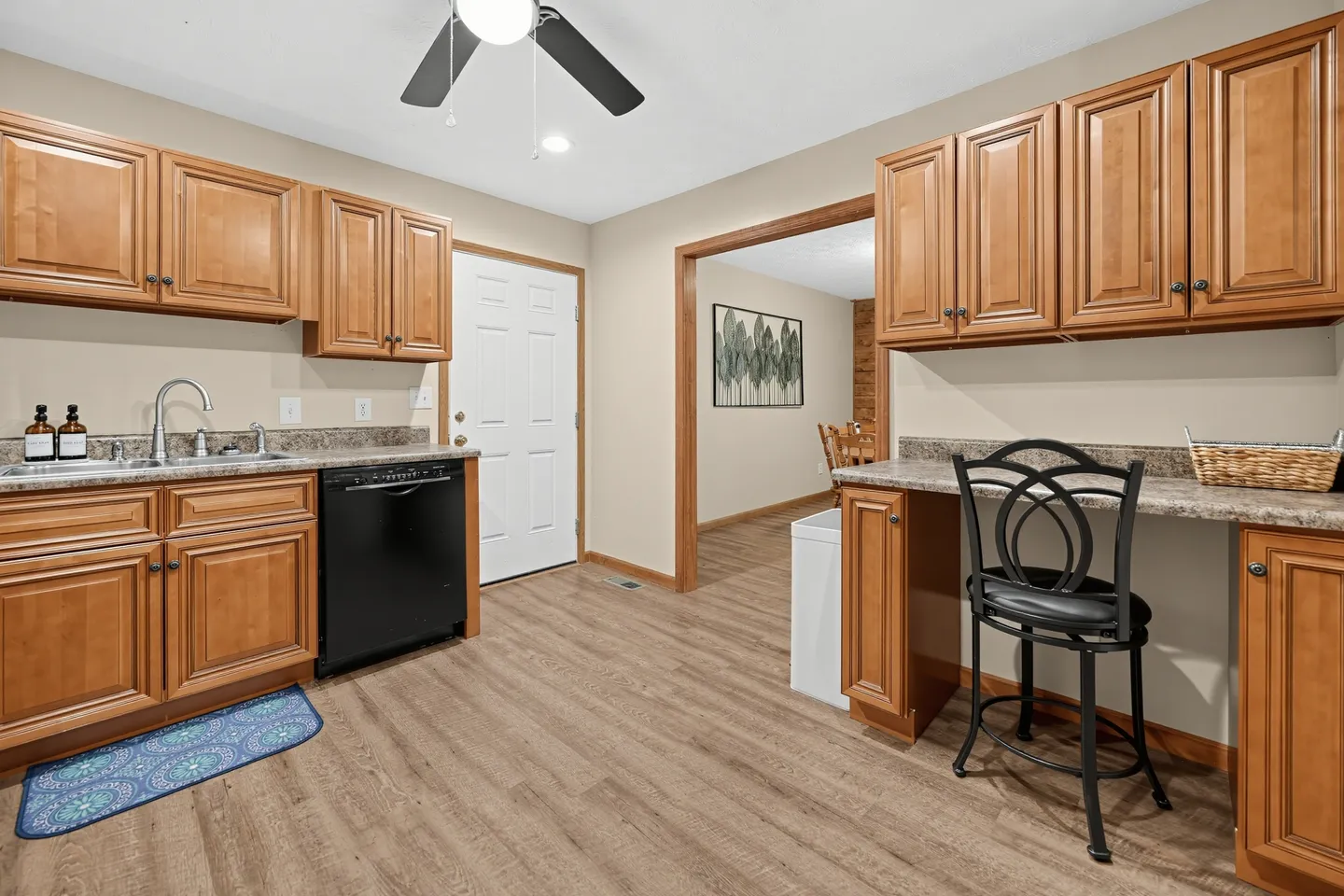 Kitchenette or utility area with wood cabinets, sink, black dishwasher, and a built-in desk counter with a black chair.