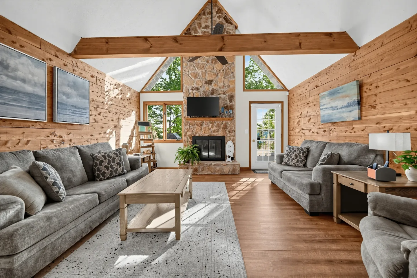 Living room with wood-paneled walls, vaulted ceiling with an exposed beam, stone fireplace, and grey sofas.