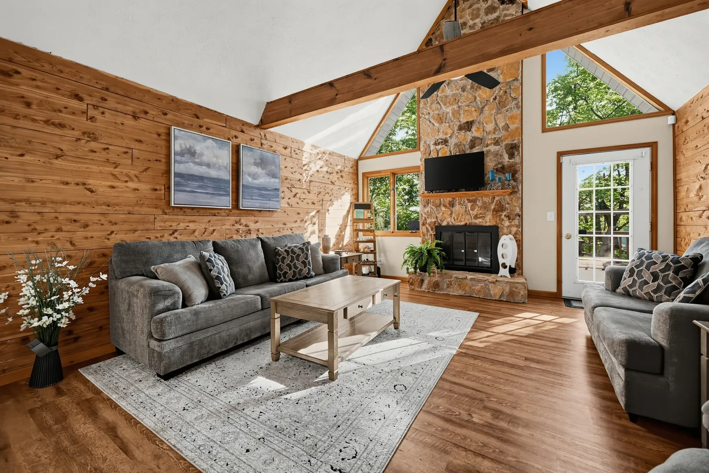 Living room with wood-paneled wall, vaulted ceiling with exposed beam, stone fireplace, and gray sofas.