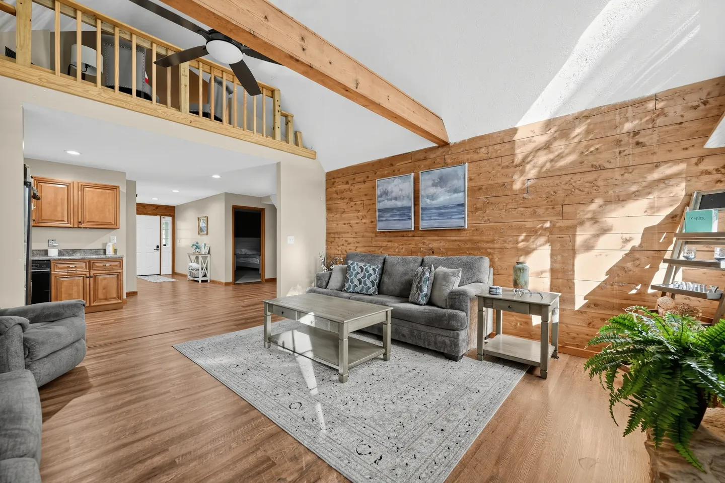 Open living area with vaulted ceiling, wood beam, loft railing above, wood-paneled accent wall, sofa, coffee table, and an adjacent kitchen in the background.