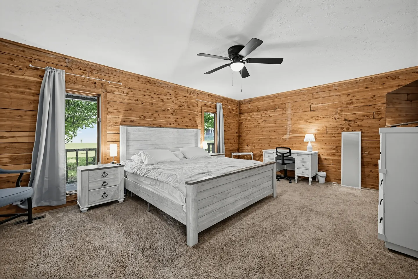 Bedroom with wood-paneled walls, carpet flooring, a light-colored wooden bed, a nightstand, and a desk with a chair.