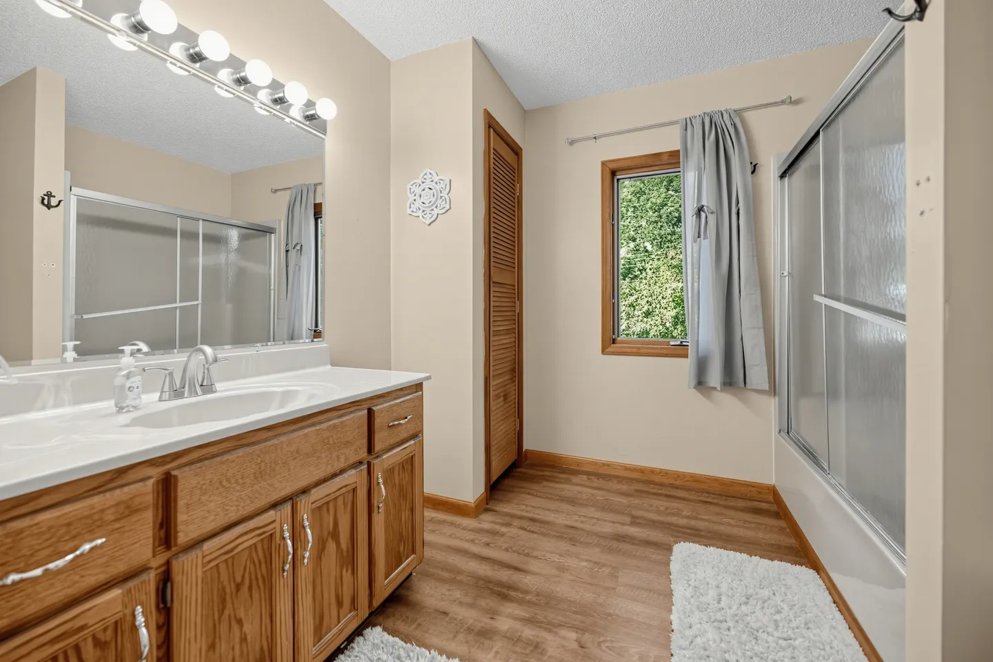 Bathroom with oak wood vanity, white countertop, lighted mirror, wood-look flooring, and a tub-shower combination with sliding glass doors.
