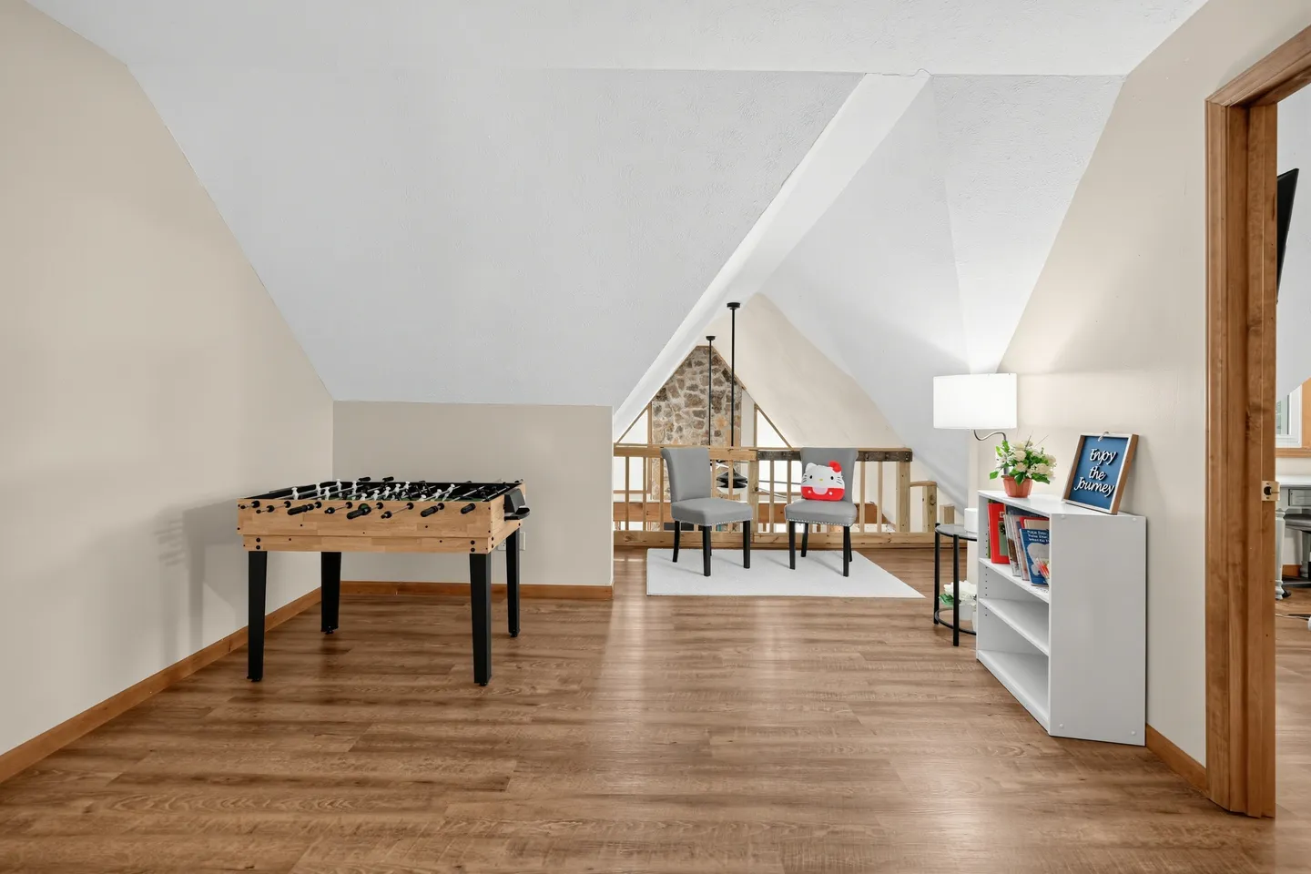 Upstairs loft game room featuring wood-style flooring, vaulted ceilings, a foosball table, a small seating area with two chairs, and a white bookcase.
