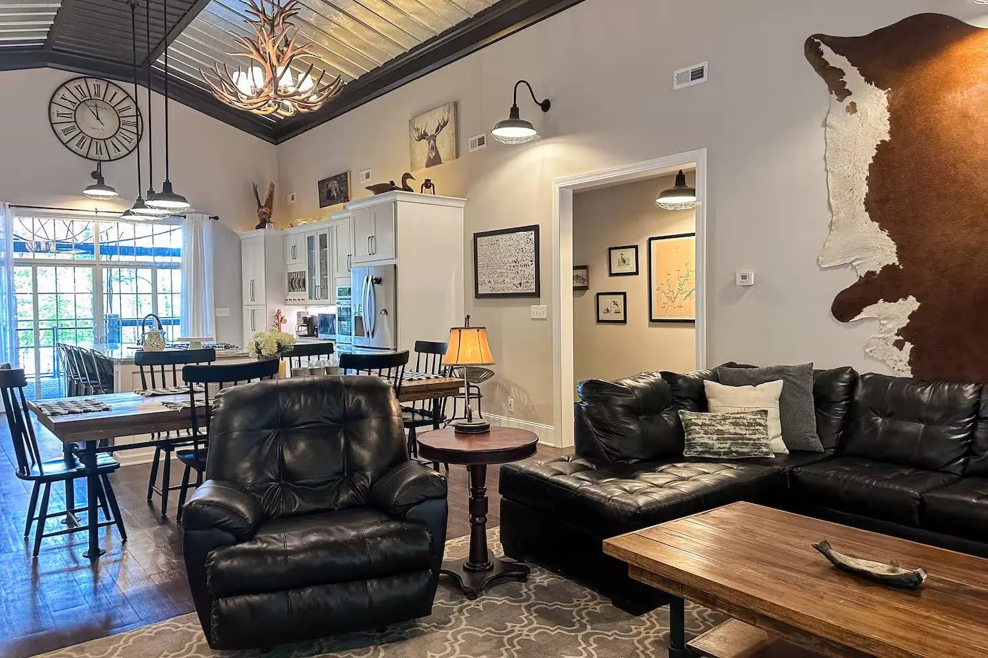 Open-concept living room and kitchen featuring high ceilings, antler chandelier, leather sectional sofa, dining table, and rustic wall decor.