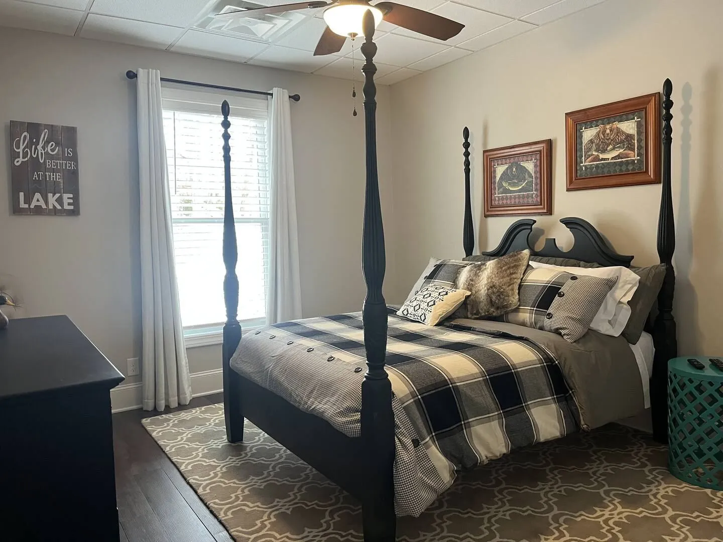 Bedroom featuring a black four-poster bed with plaid bedding, patterned area rug, and window with white curtains.