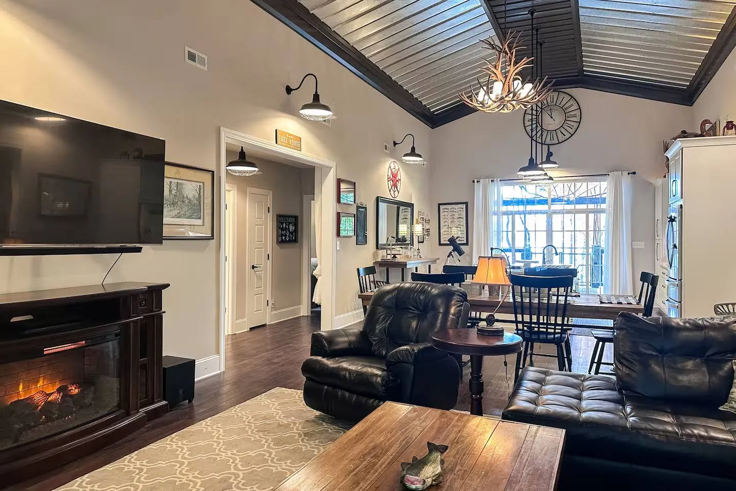 Open-concept living room with a leather recliner, fireplace, wall-mounted television, and high vaulted ceiling leading toward a dining area.