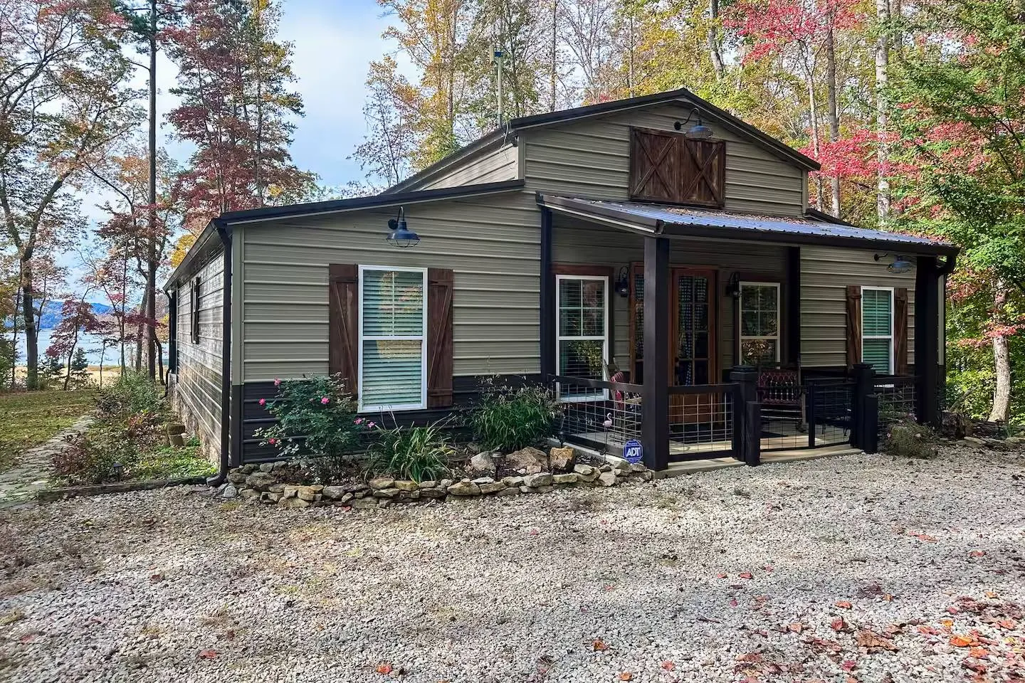 Exterior of a metal siding cabin with a front porch, gravel driveway, and trees with water in the background.