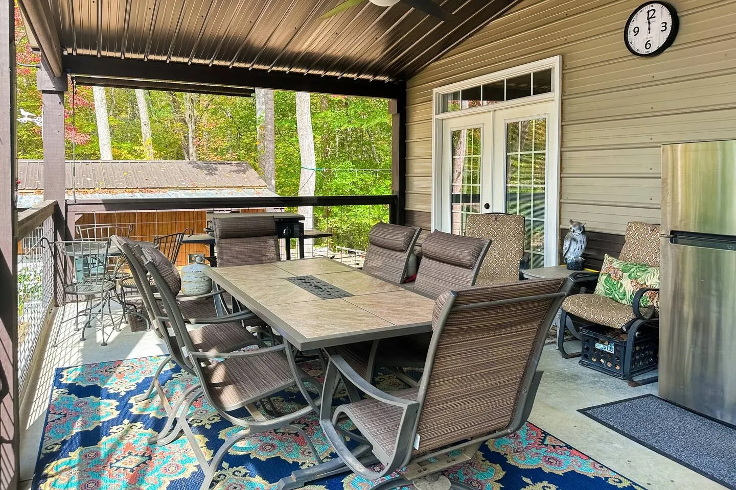 Covered patio featuring an outdoor dining table with chairs, an outdoor rug, refrigerator, and grill overlooking a wooded yard.