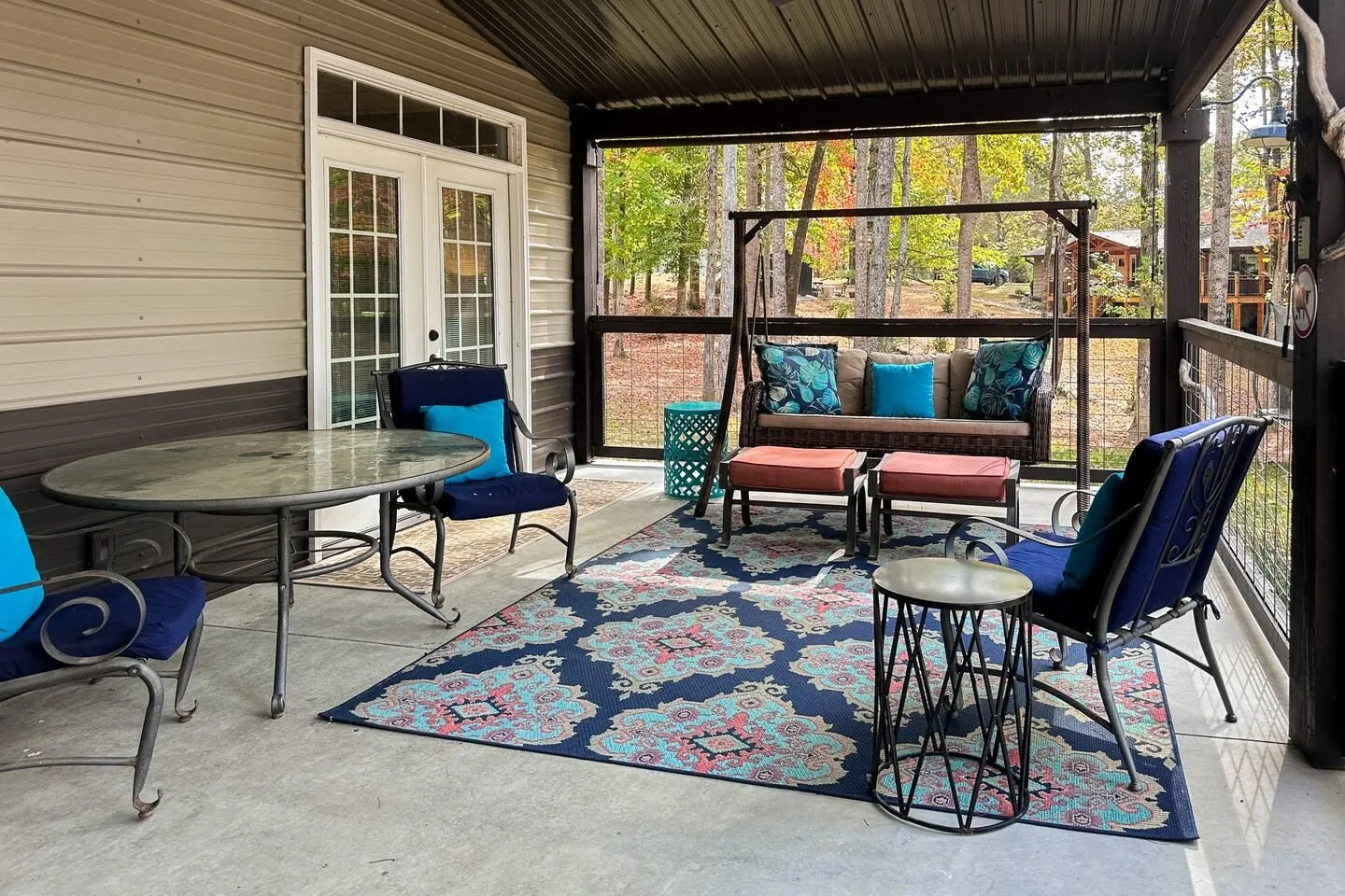 A covered patio with a patterned outdoor rug, cushioned chairs, a swing bench, and a dining table, looking out onto trees.