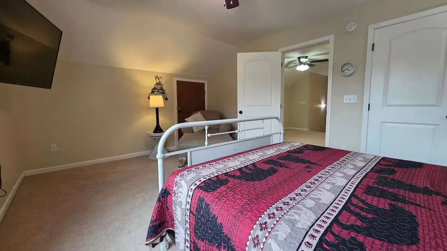 Bedroom with vaulted ceiling, carpeted floor, metal bed frame with red patterned quilt, armchair, and open doorway.