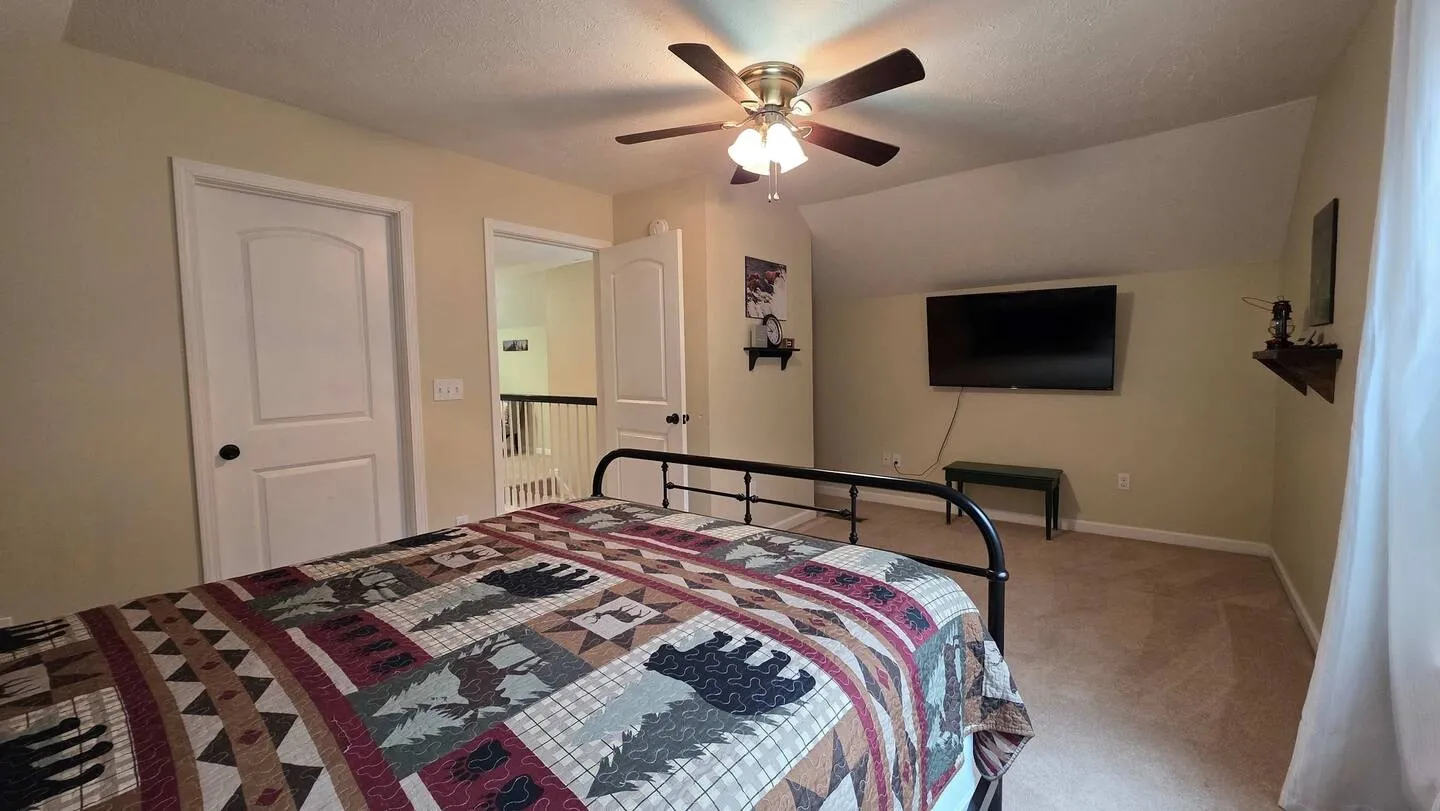 Bedroom with a patterned quilt on a black metal bed frame, a wall-mounted flat-screen TV, and a lighted ceiling fan.