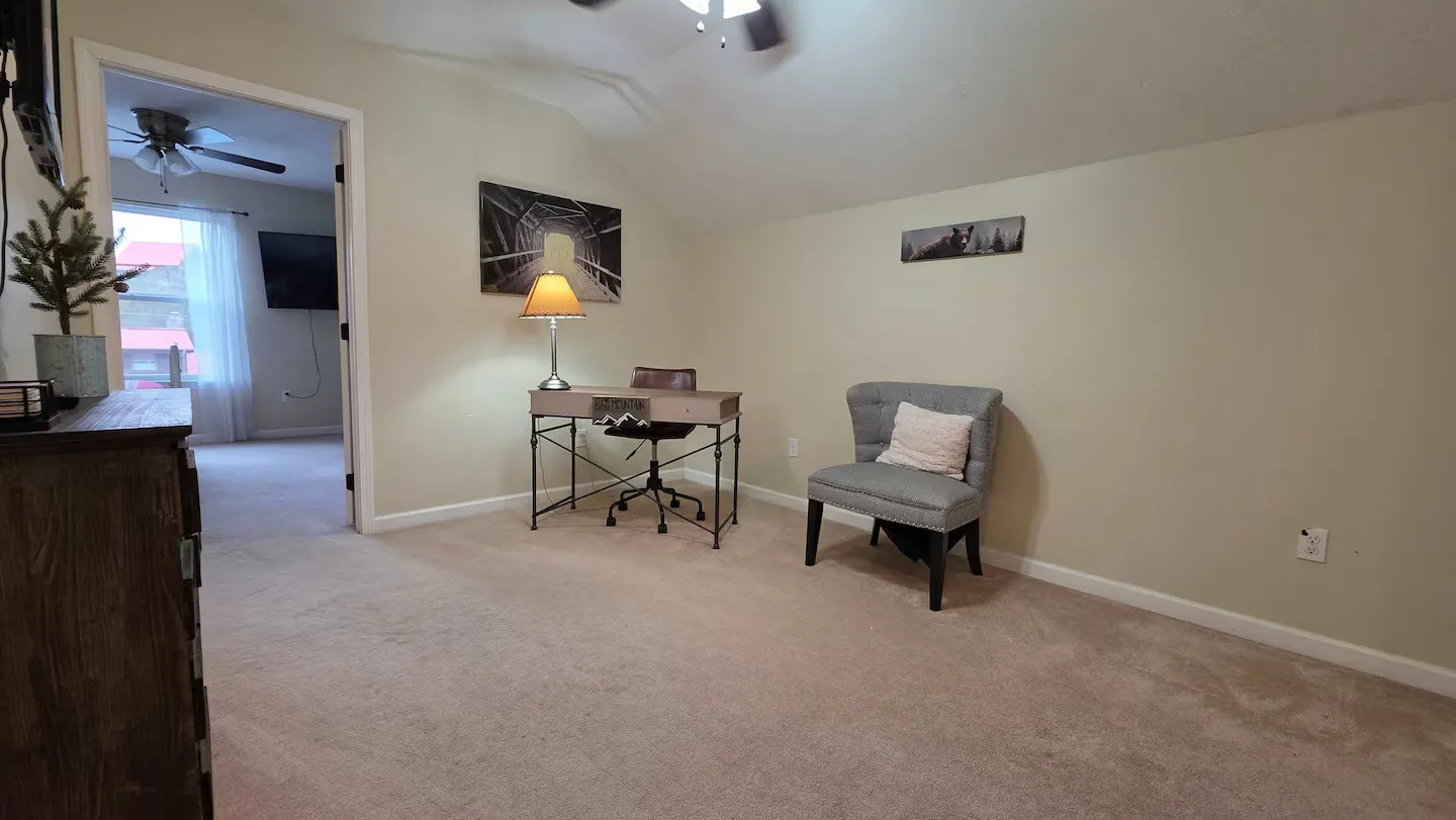 A carpeted upstairs workspace area featuring a desk, office chair, upholstered armchair, and an open doorway leading to another room.