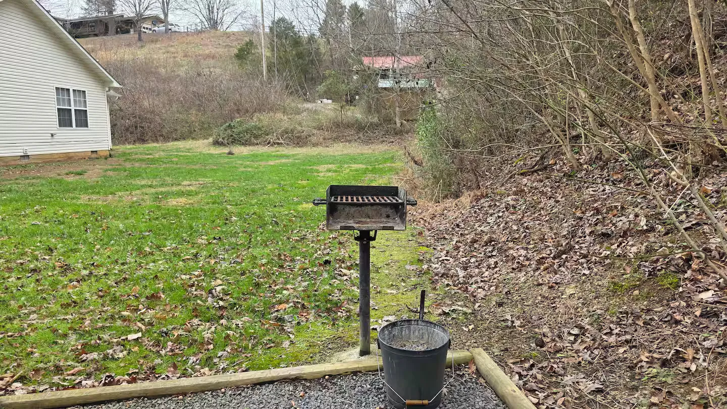 Park-style charcoal grill mounted on a post in a grassy yard beside a wooded slope.