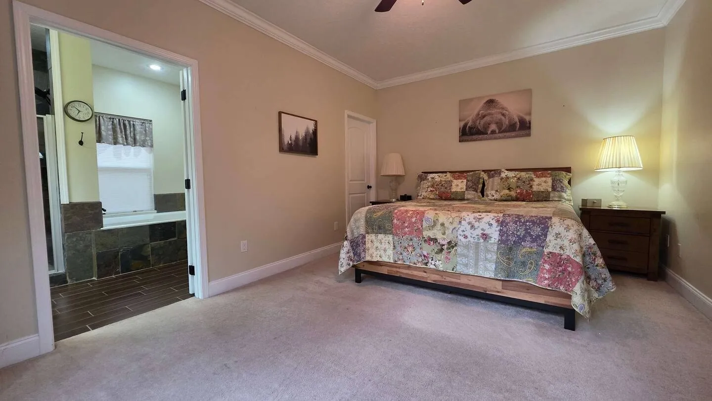 Bedroom with carpet, a patterned quilt on the bed, nightstands with lamps, and an open doorway leading to an en-suite bathroom.