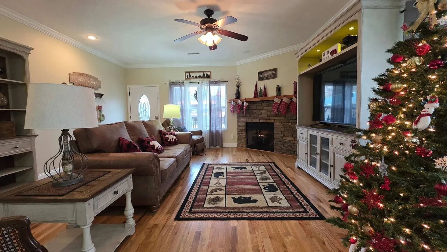 Living room with wood flooring, brown sofa, stone corner fireplace, large entertainment center, and decorated Christmas tree.