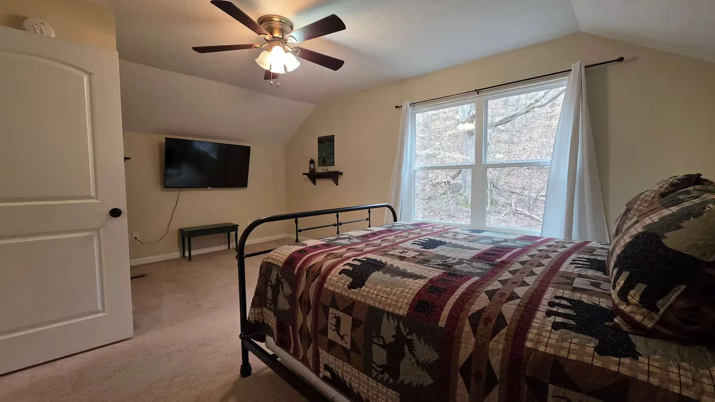 Bedroom with a metal bed frame, patterned wildlife quilt, wall-mounted television, and a window with wooded views.