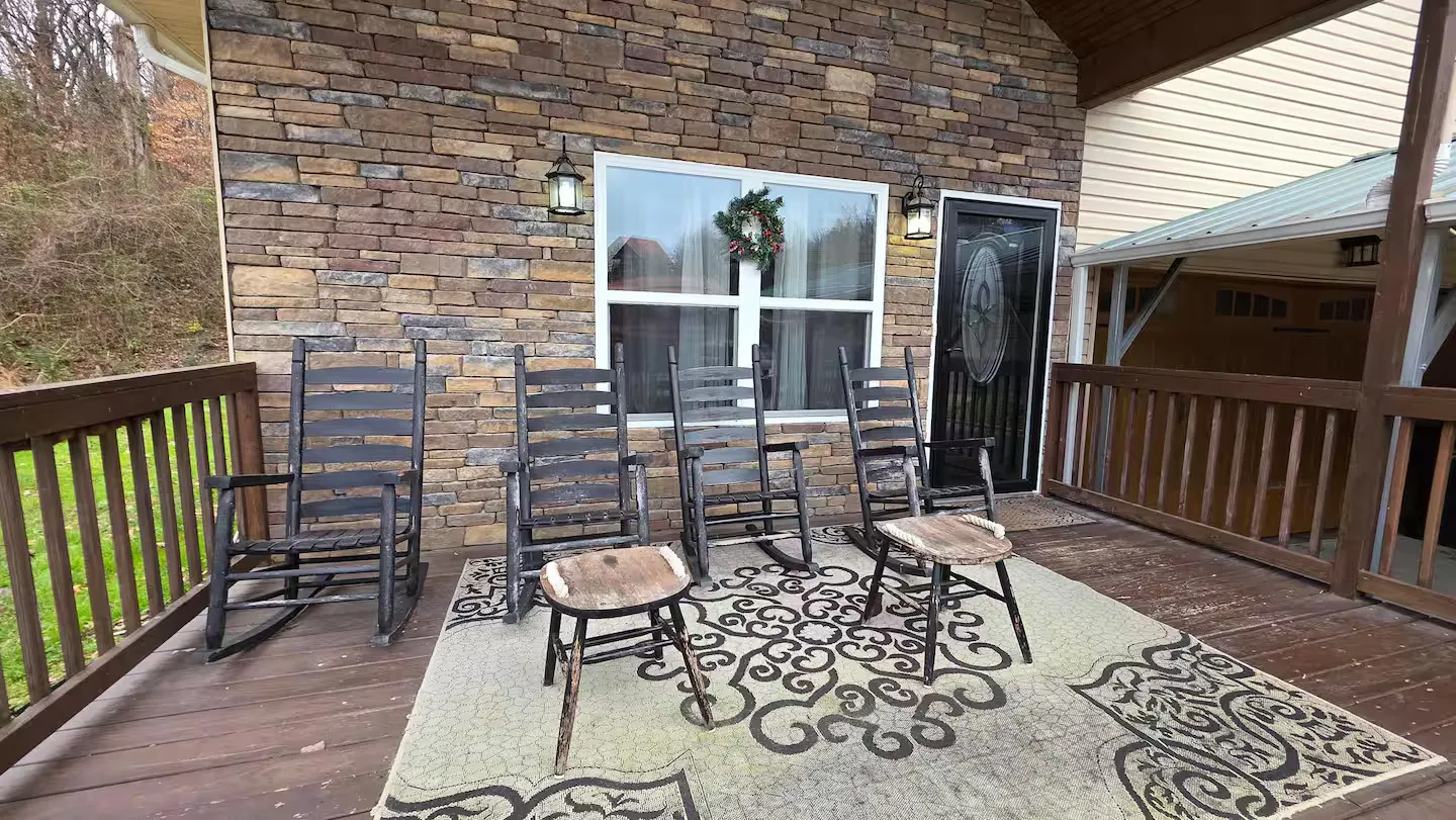 Porch deck with four wooden rocking chairs, two small footstools, an outdoor rug, stone siding, and an entry door.