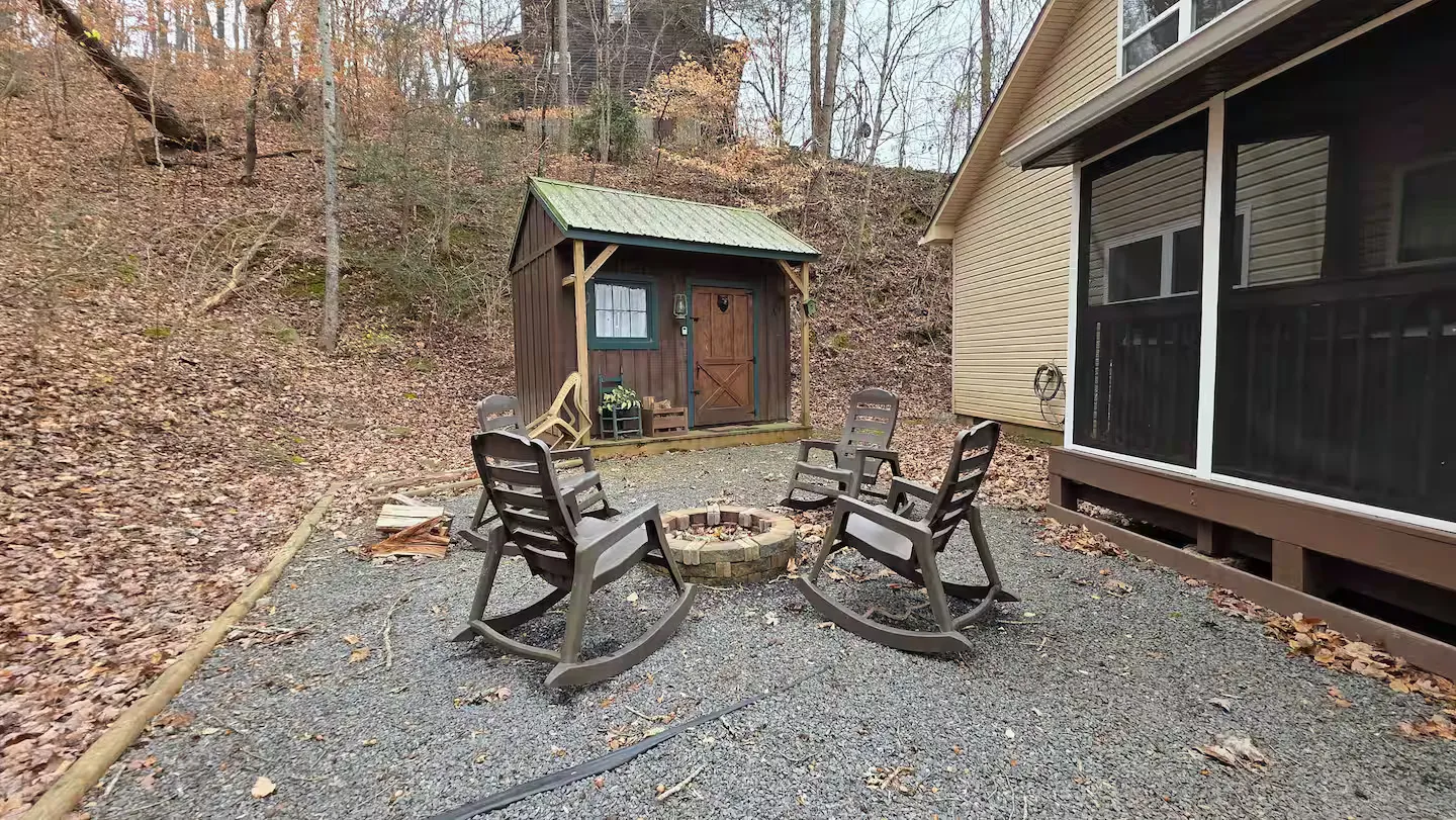 Outdoor gravel patio featuring a stone fire pit surrounded by rocking chairs next to a small wooden shed and house.