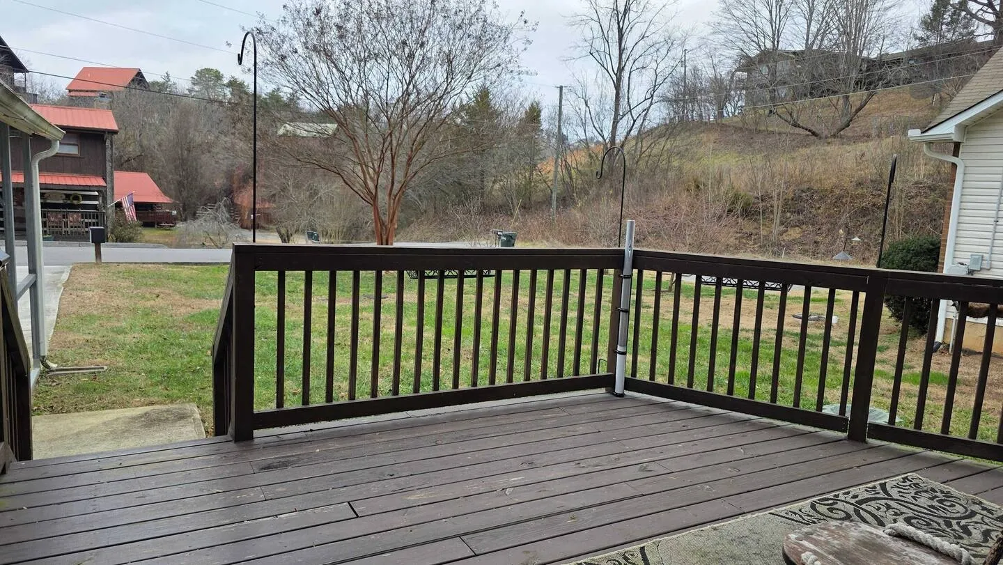 Outdoor wooden deck with dark brown railings overlooking a grassy yard, trees, and distant cabins under an overcast sky.