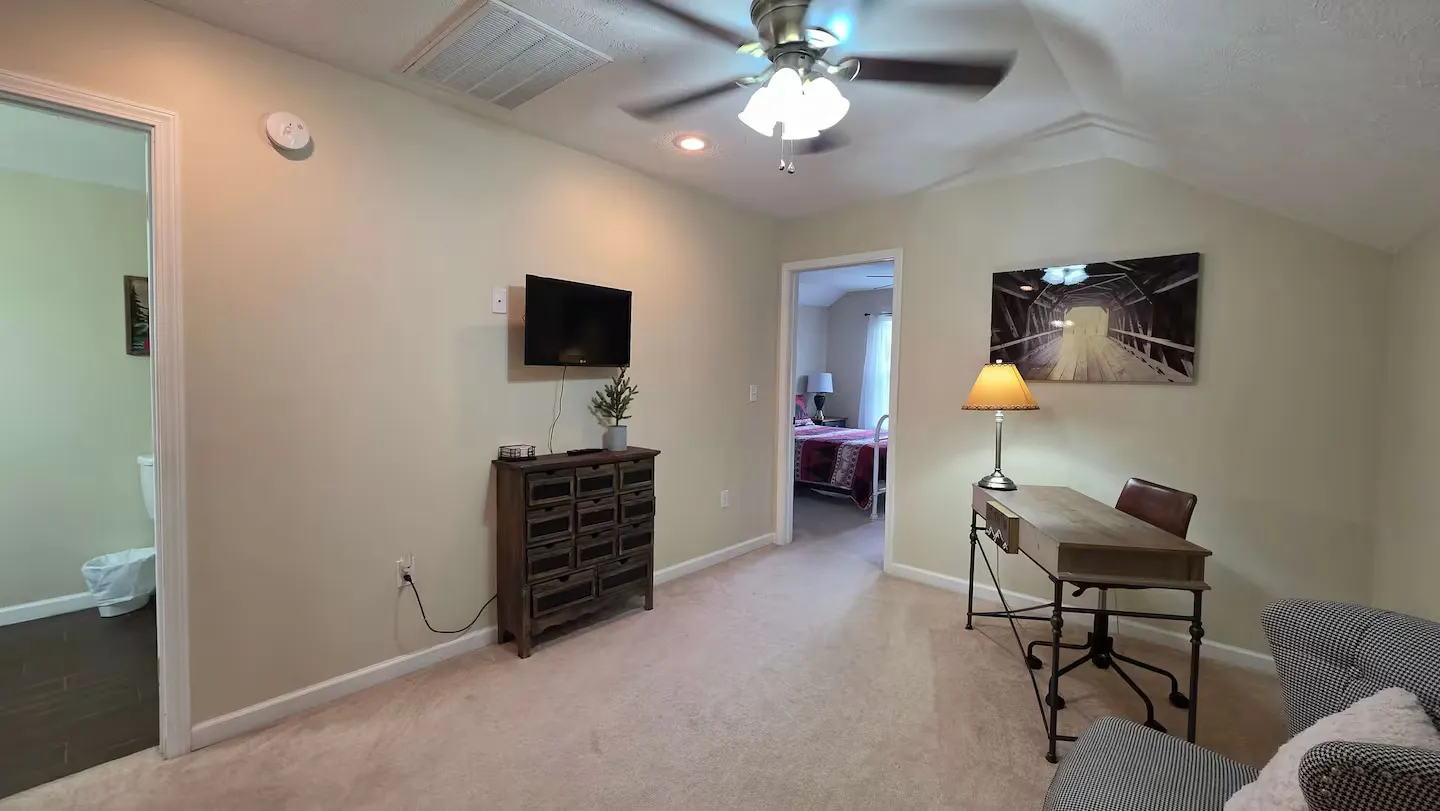 A carpeted upstairs sitting room with a work desk, television on the wall, and views into an adjacent bathroom and bedroom.