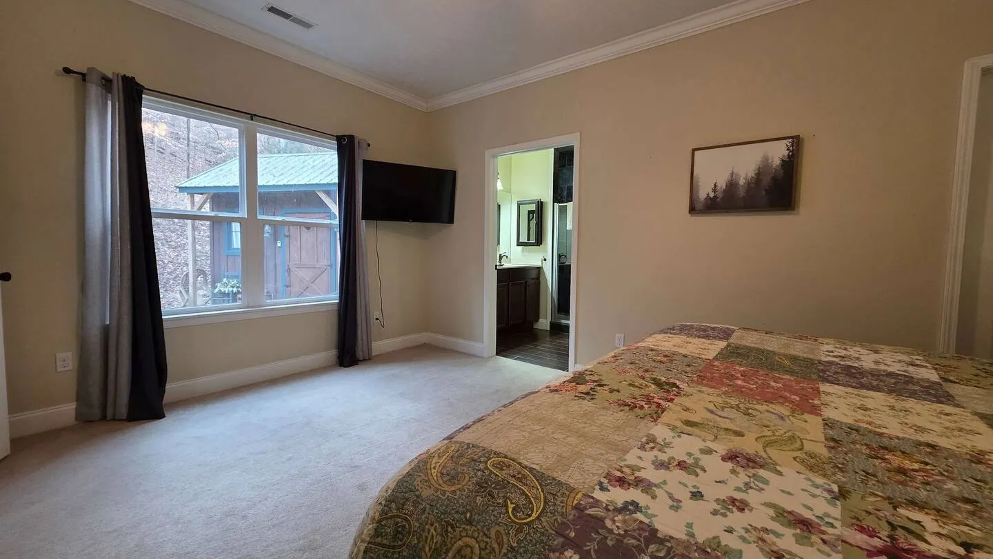 Bedroom with carpet, a large bed with a patterned quilt, a mounted TV, a window looking outside, and a doorway to an en suite bathroom.