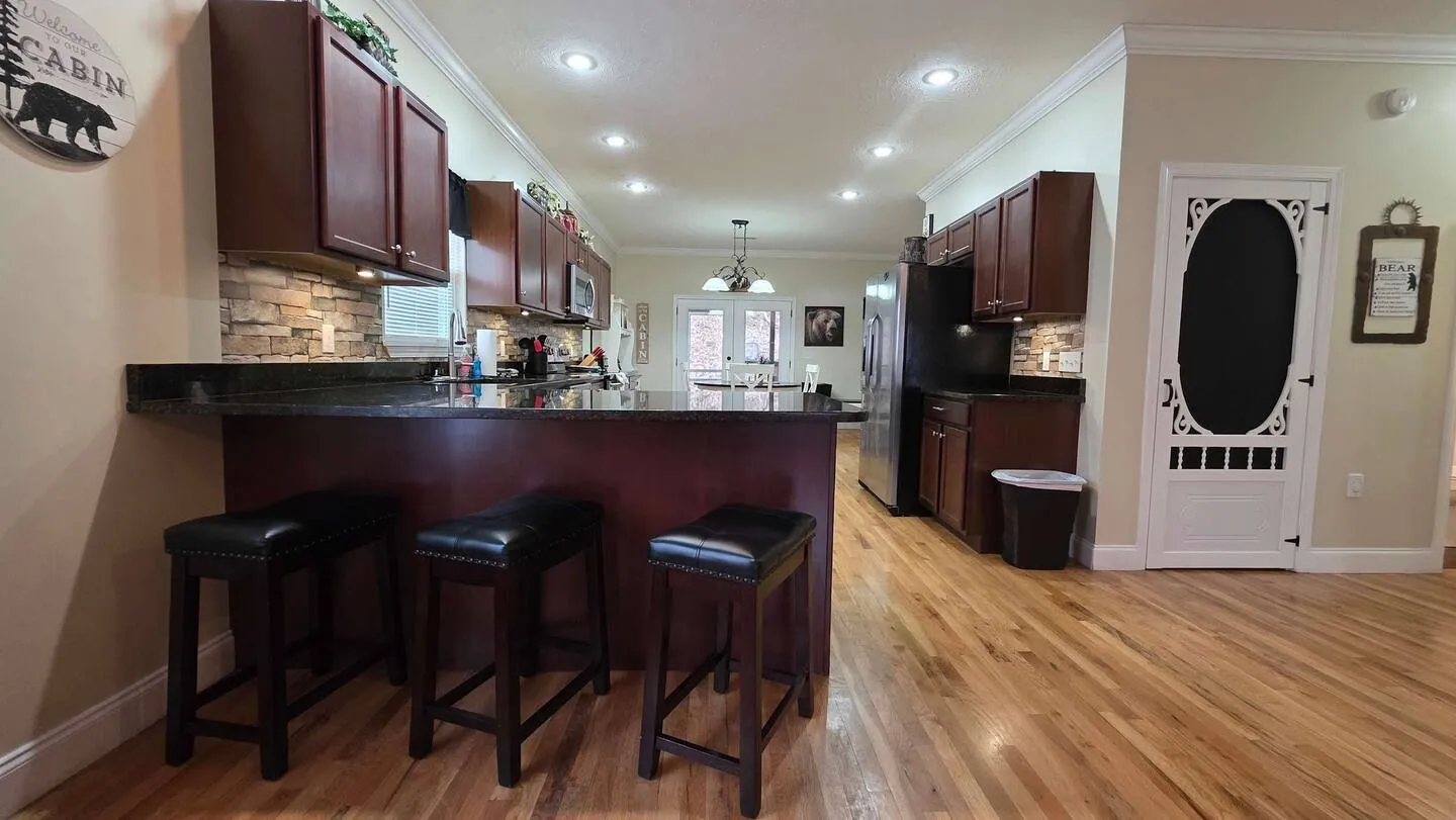 Kitchen counter with three dark backless barstools, dark wood cabinetry, and a view into the open dining and living space.