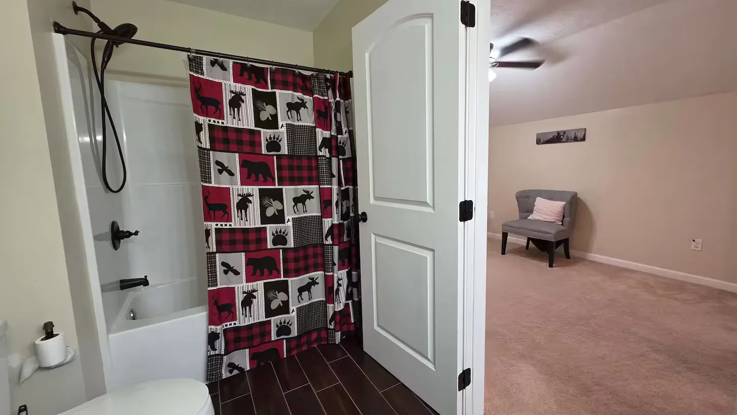 Bathroom with shower-tub combo and patterned curtain opening to a carpeted room with an accent chair.