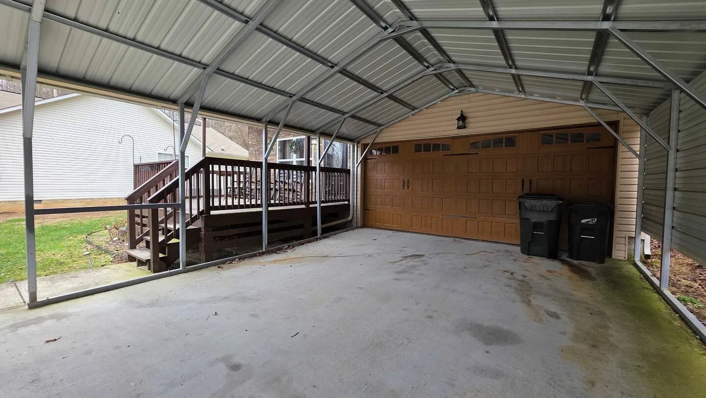 A covered metal carport on a concrete pad leading to a wooden garage door with outdoor steps and deck visible to the side.
