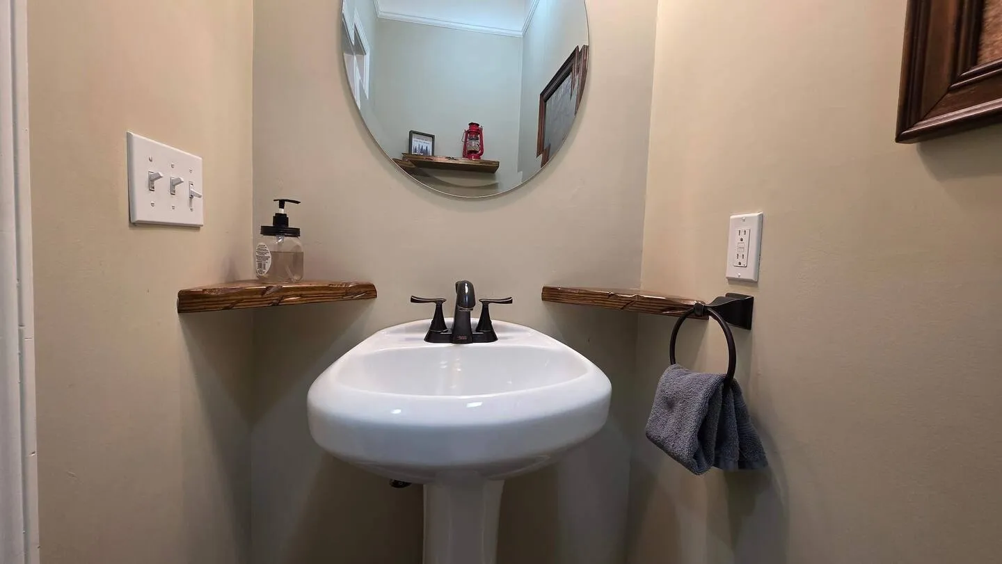 Powder room with a white pedestal sink, dark metal faucet, circular mirror, and small wooden corner shelves.