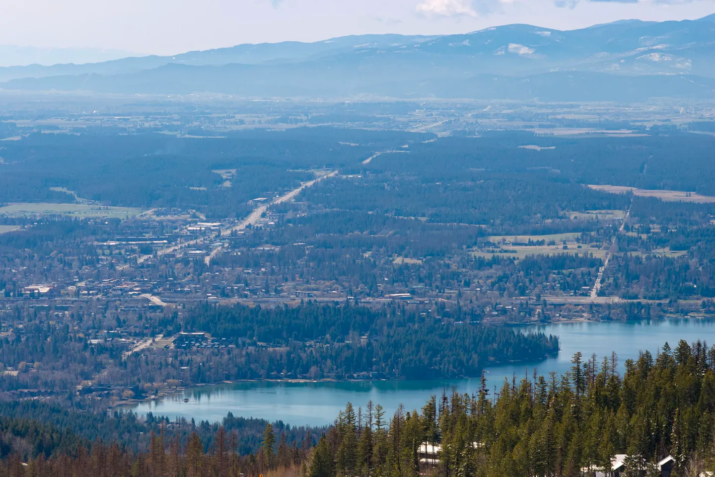 Elevated scenic overlook of a lake surrounded by dense evergreen forests and distant mountains under a blue sky.