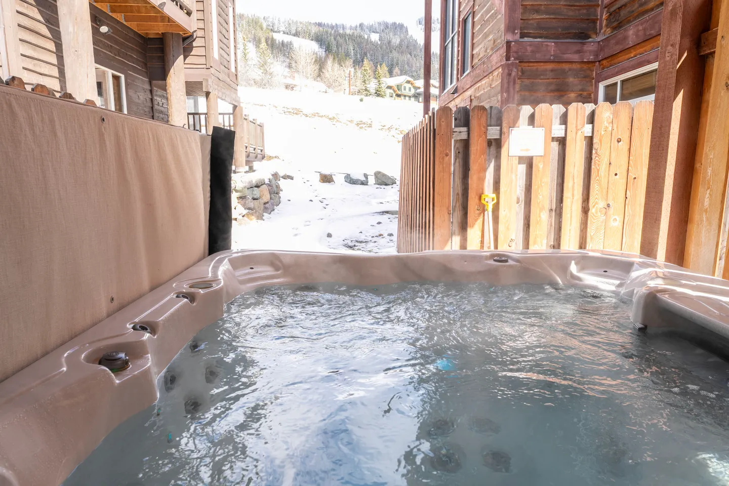 Outdoor hot tub filled with bubbling water looking out toward a snowy hillside and neighboring wooden cabins.