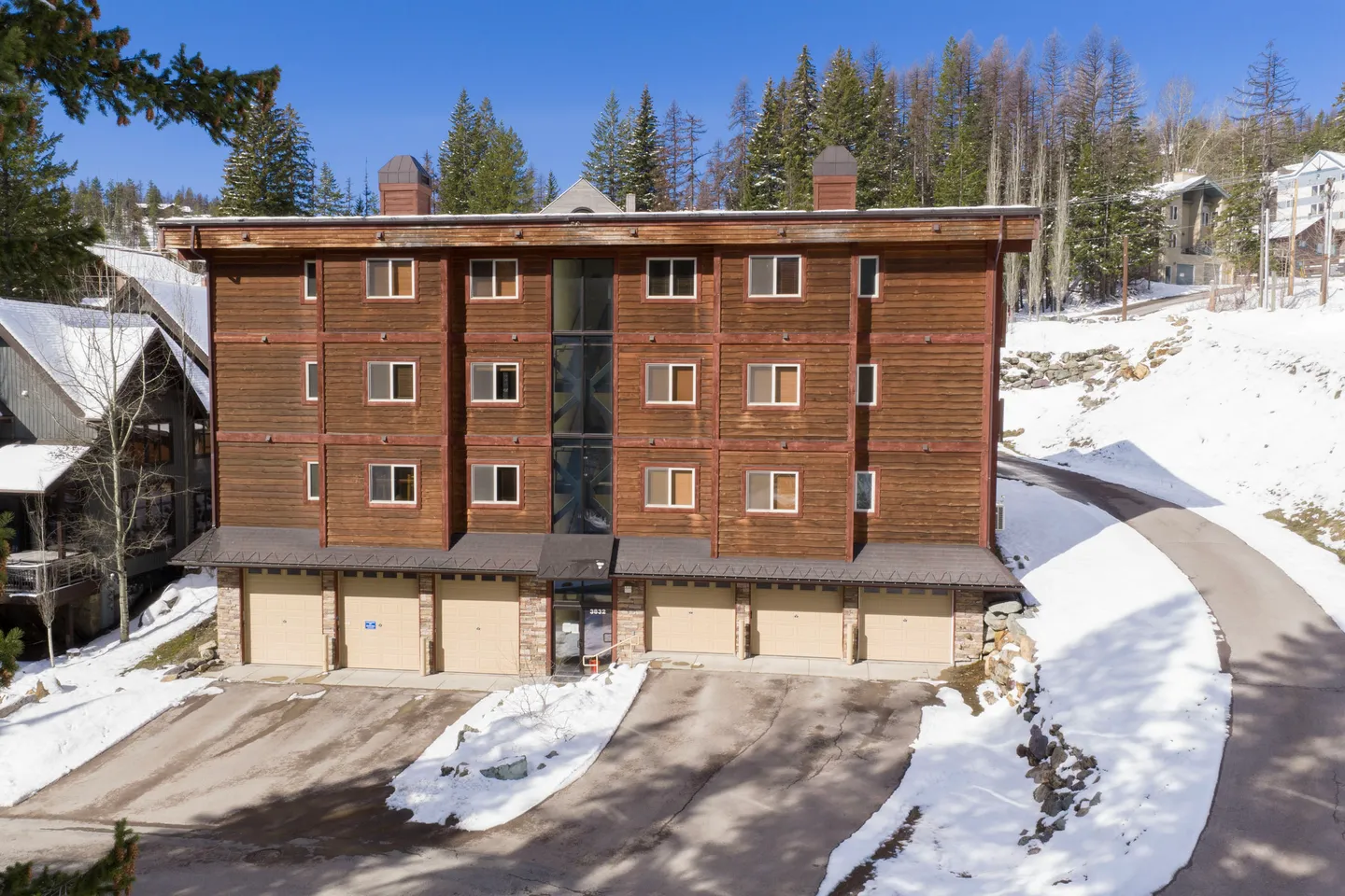 Exterior view of a multi-story wood-sided building with ground-level garages surrounded by snow-covered ground and pine trees.
