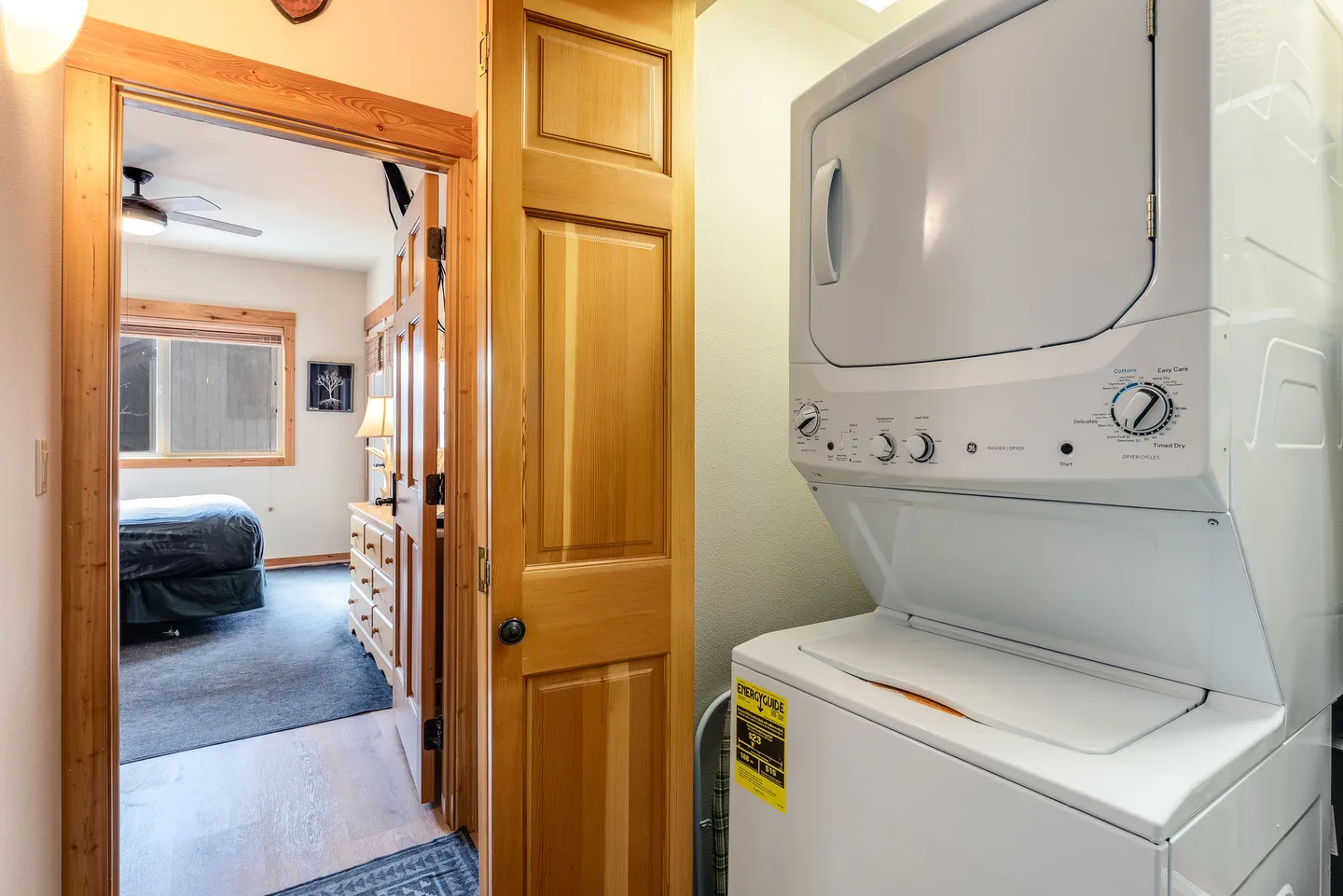 A stacked white washer and dryer in a laundry closet looking into an adjacent bedroom.