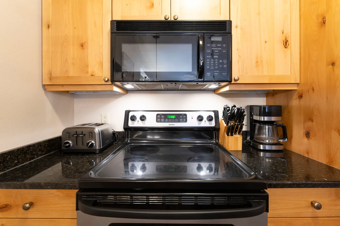 Close-up of a kitchen cooking area with a glass-top stove, over-the-range microwave, toaster, knife block, and coffee maker on dark speckled countertops surrounded by light wood cabinetry.