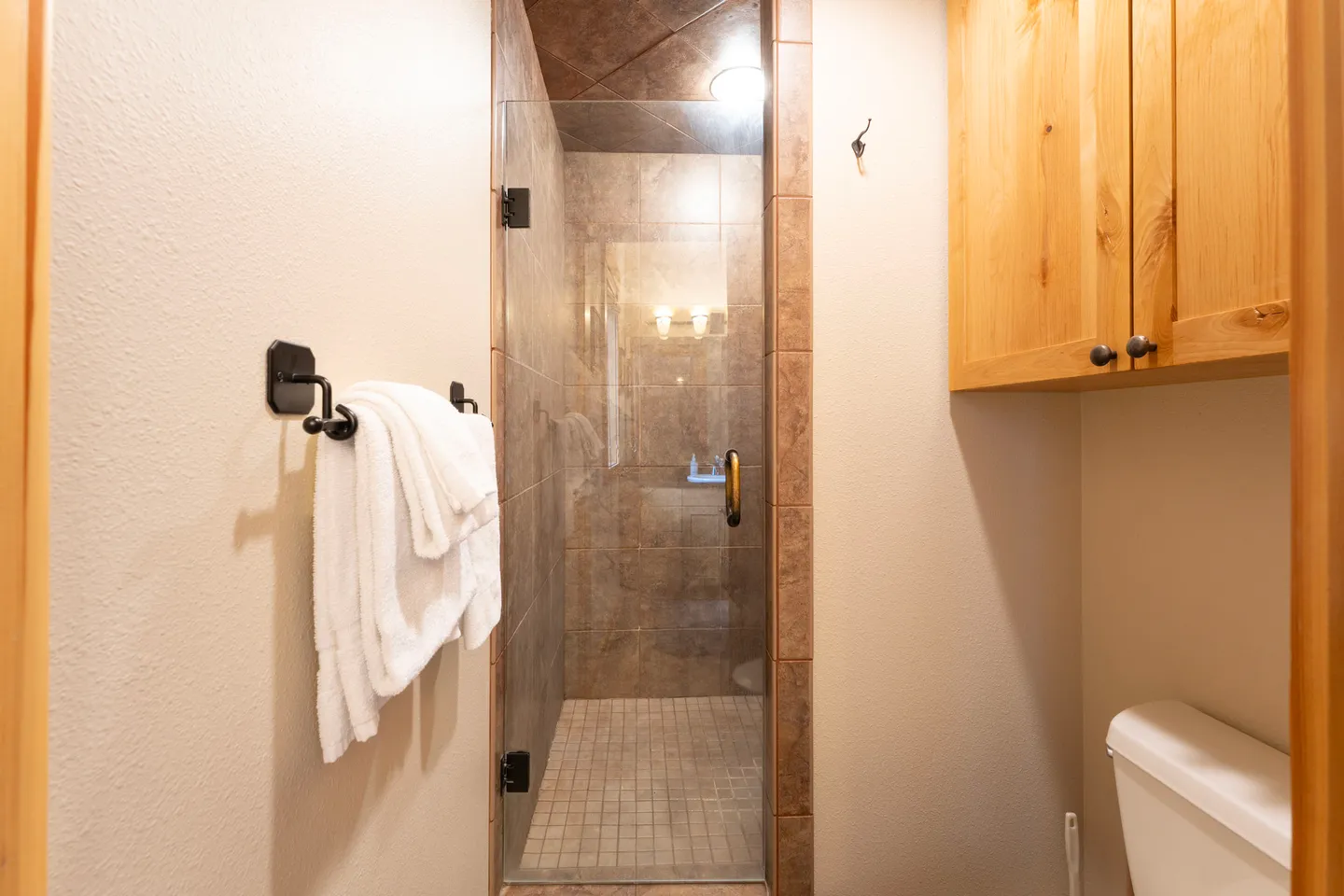 Bathroom showing a walk-in shower with a glass door, light brown tile, white towels on a black rack, and wood upper cabinets over a toilet.