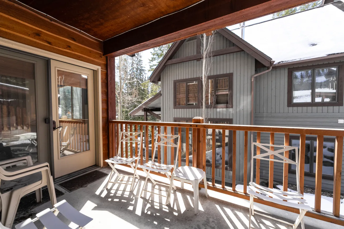 Covered outdoor balcony with wooden railings and white chairs looking out at neighboring cabins and snow.