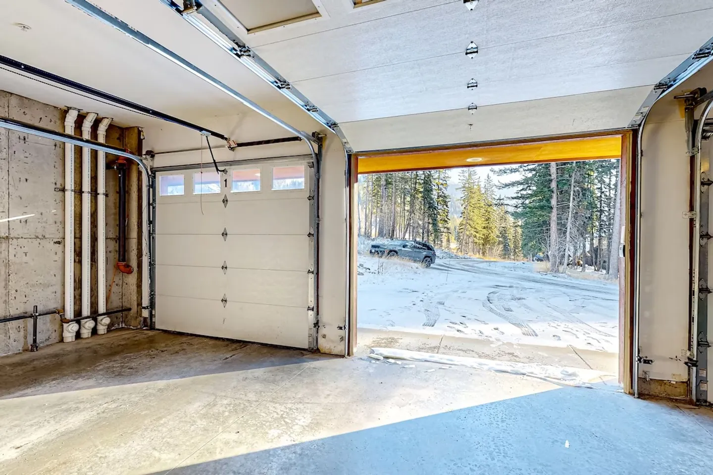 Interior view of a concrete-floored garage looking out an open bay door onto a snowy driveway surrounded by pine trees.