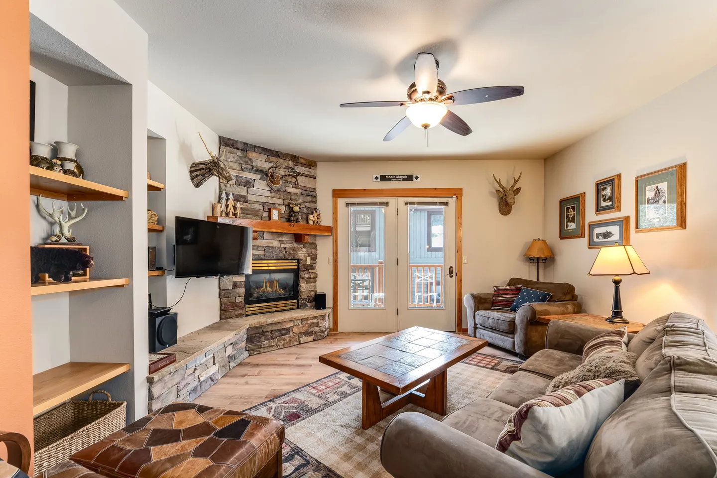 Living room with a stone corner fireplace, mounted TV, sectional sofa, armchair, and glass doors leading outside.