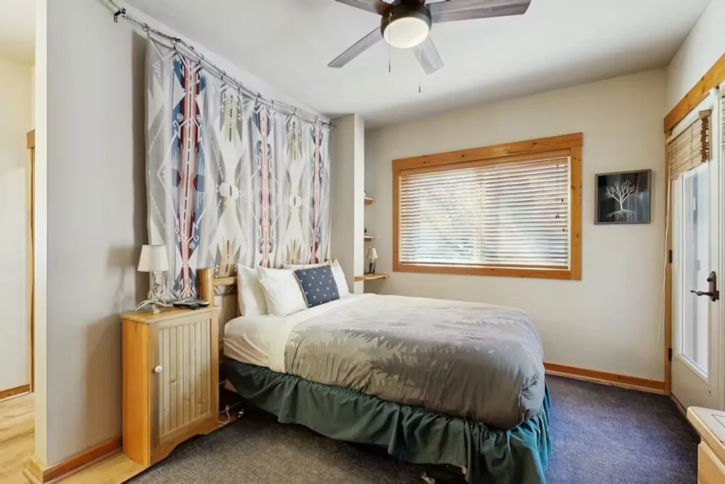 Bedroom featuring a made bed with a patterned tapestry headboard, a ceiling fan, carpet, and a wood-trimmed window with horizontal blinds.
