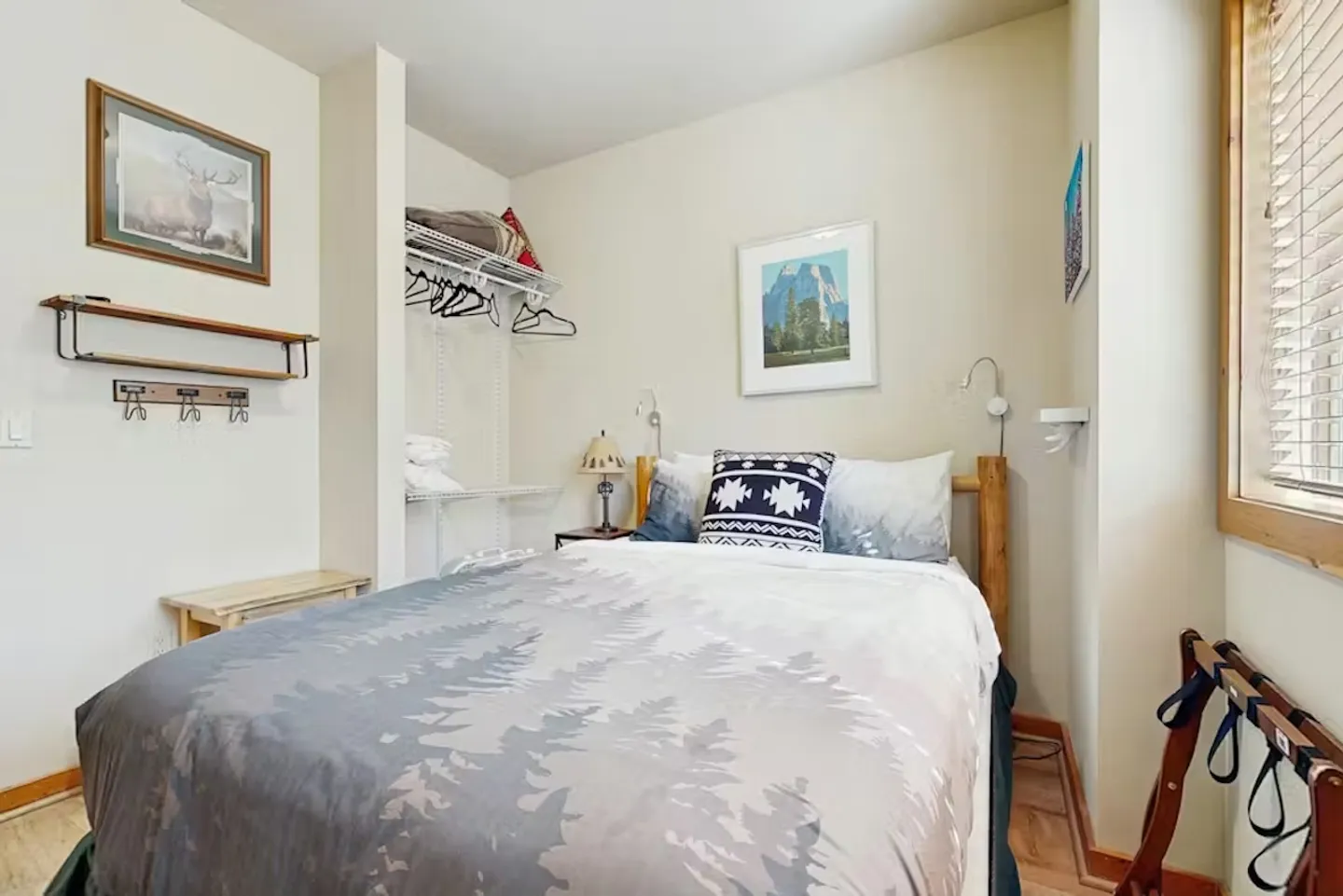 Bedroom featuring a made bed with a nature-themed comforter, open wire closet shelving, and a luggage rack by a window with blinds.