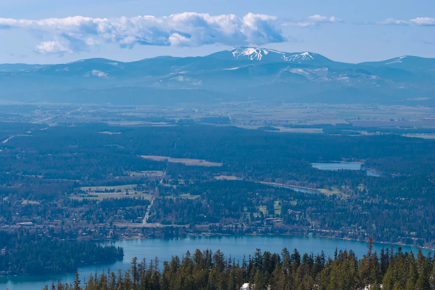 Elevated scenic view overlooking a lake, evergreen forests, and distant snow-capped mountains beneath a blue sky.