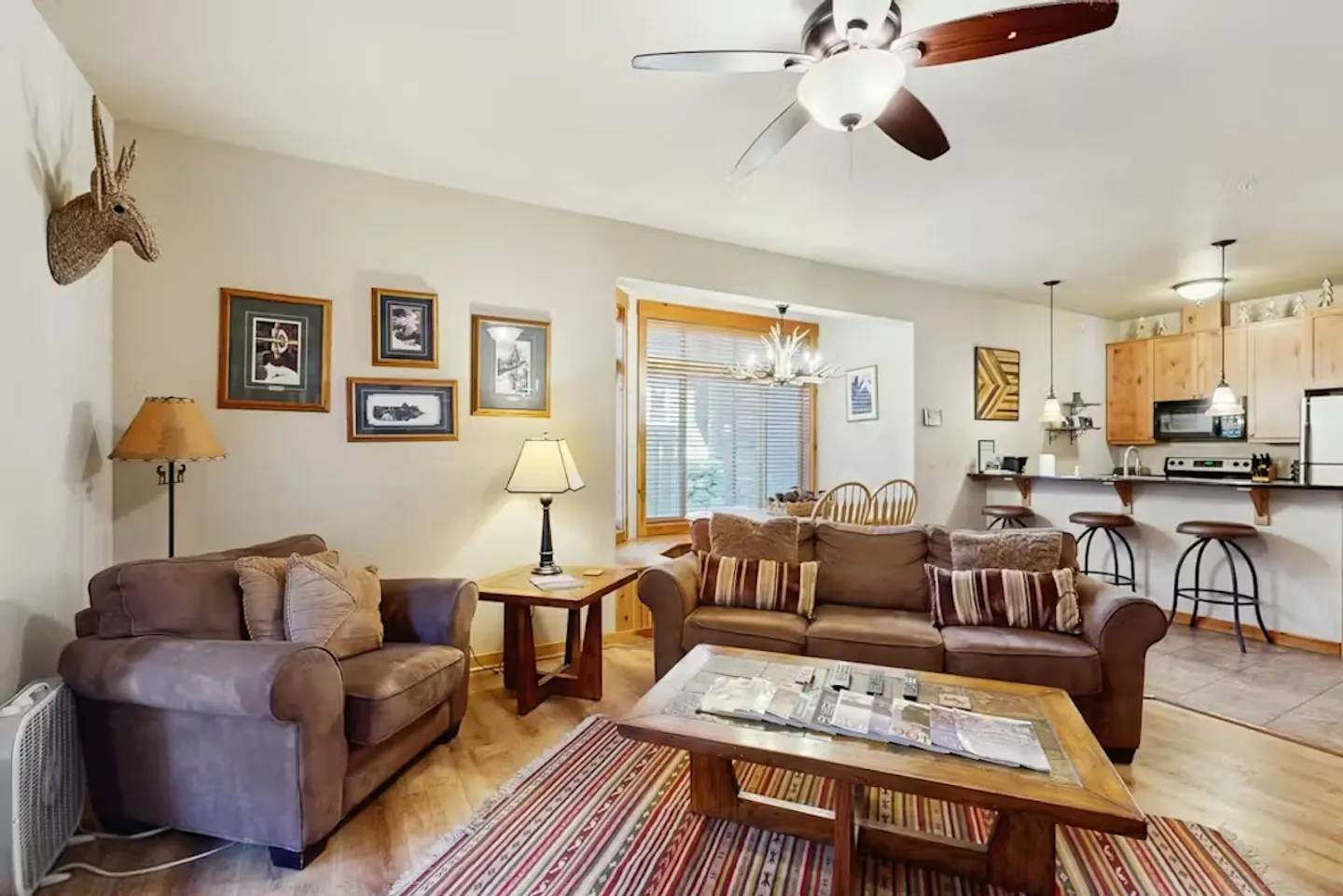 Living area with a brown sofa, armchair, coffee table, ceiling fan, and view into the kitchen and dining nook.