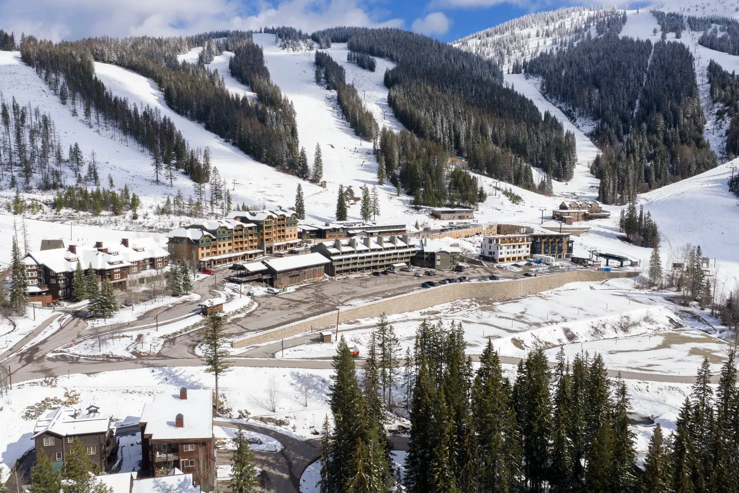 Elevated view of a mountain ski village surrounded by snow-covered slopes, pine trees, and ski runs under a clear blue sky.