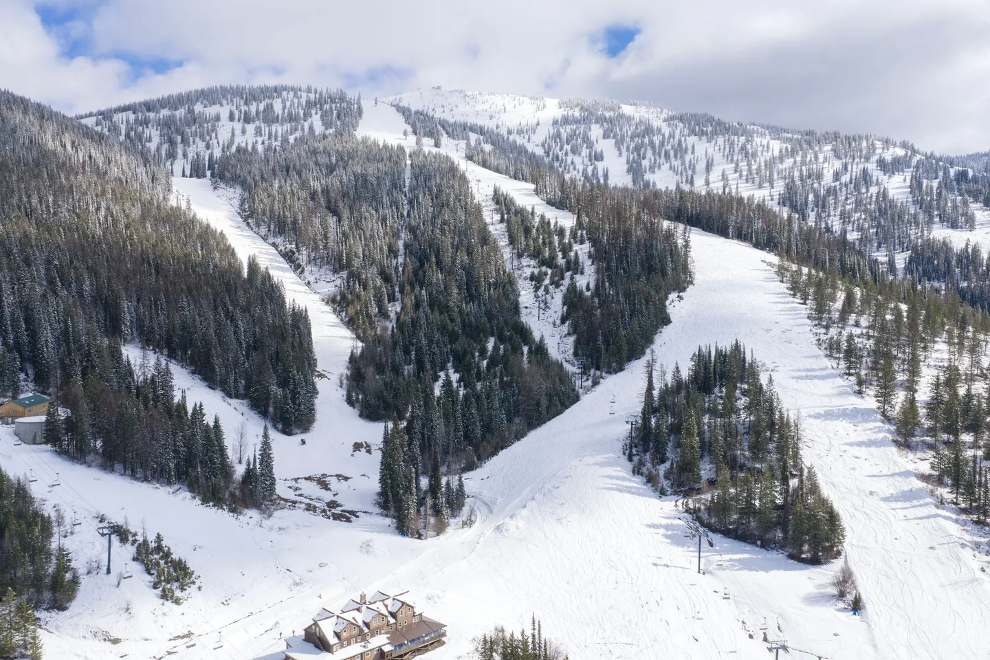 Aerial view of a snow-covered mountain with ski runs, evergreen trees, chairlifts, and a lodge at the base.