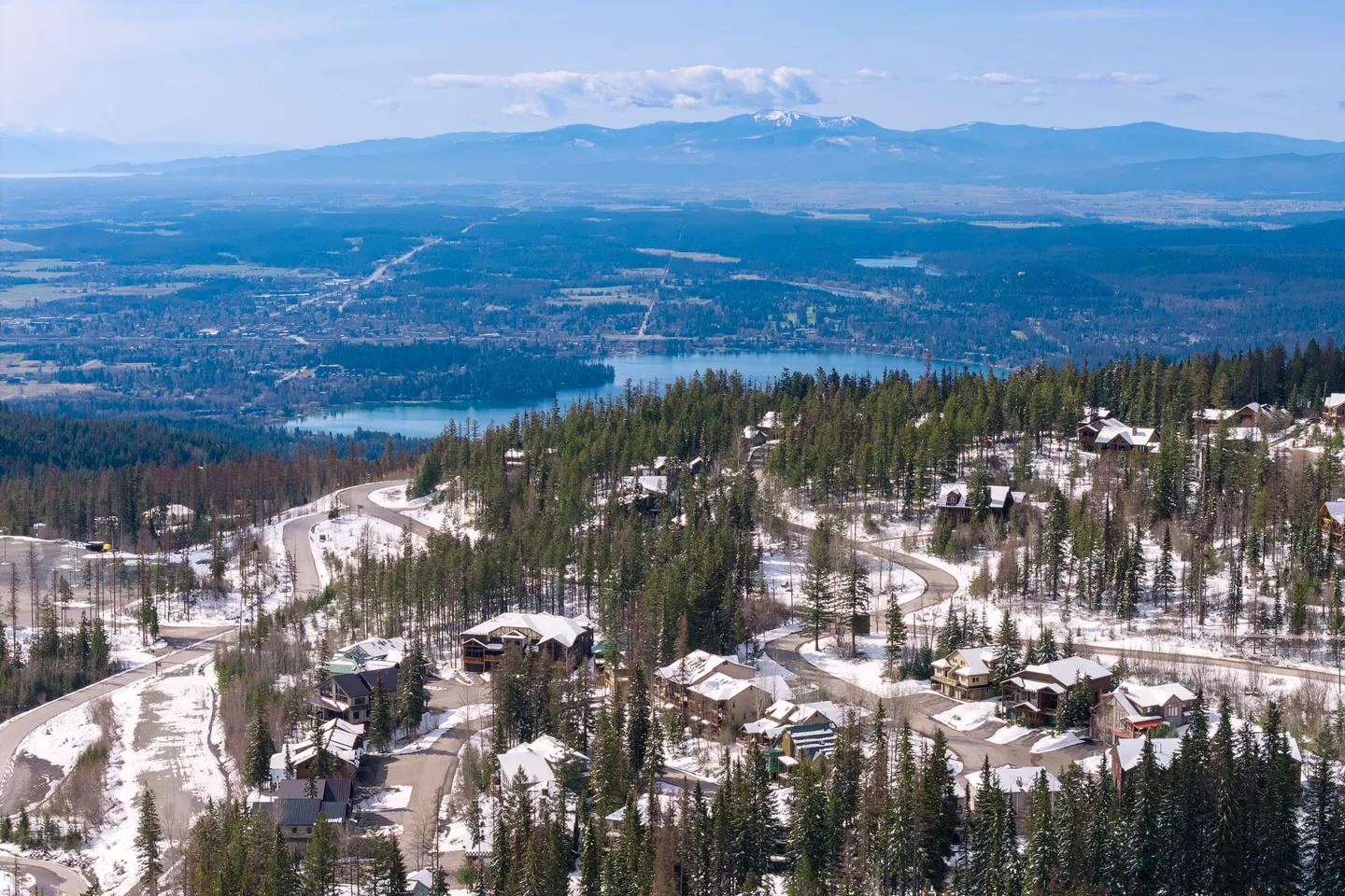 Aerial view of a mountainside neighborhood with scattered snow, evergreen trees, and a lake in the distant valley.