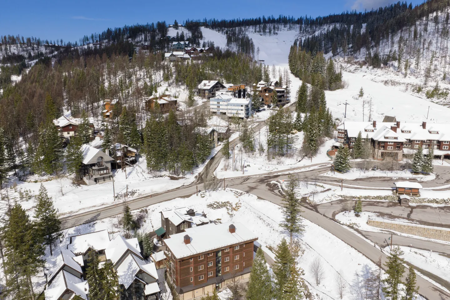 Aerial view of a snowy mountain village with roads, lodges, evergreen trees, and ski slopes in the background.