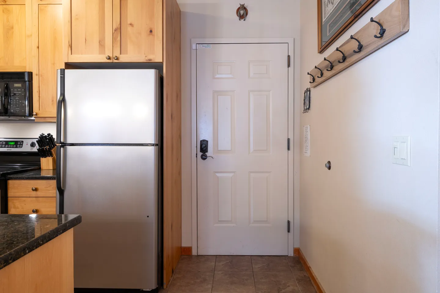Entryway with a white six-panel front door, coat rack on the wall, and adjacent kitchen with stainless steel refrigerator and wood cabinetry.