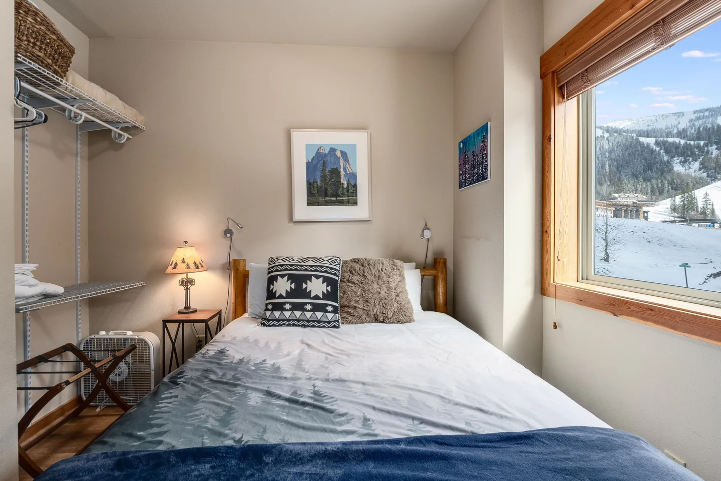 Bedroom with a bed dressed in forest-themed bedding, a wire shelving system on the left, and a window with a snowy mountain view.